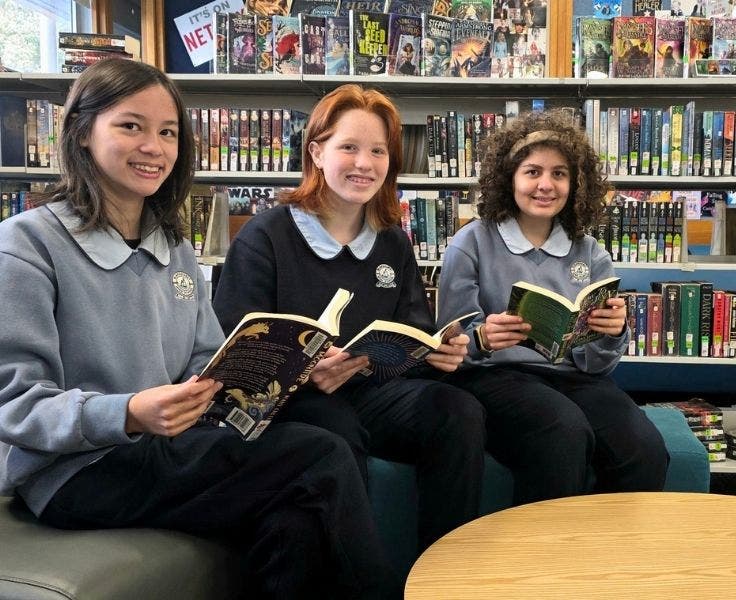 An image of three students in the library holding a book sitting amongst the library shelves smiling at the camera.